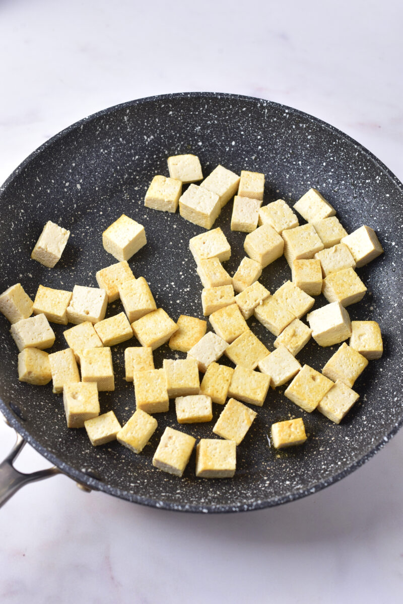 Cubed tofu pieces cooking in a black nonstick frying pan on a light-colored surface.