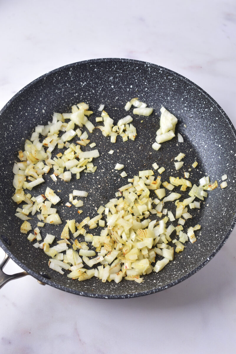 Chopped onions being sautéed in a black non-stick frying pan on a light-coloured surface.