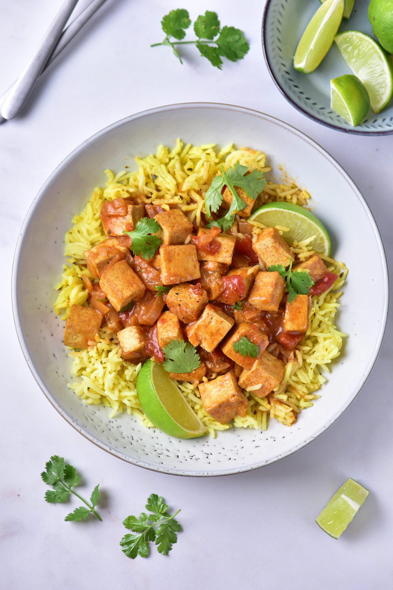 A bowl of yellow rice topped with flavorful Indian tofu curry, garnished with fresh cilantro and lime wedges. Extra cilantro and lime pieces are on the table beside the bowl.