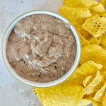 A bowl of creamy black bean dip sits next to a pile of yellow tortilla chips on a light surface.
