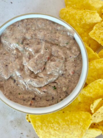 A bowl of creamy black bean dip sits next to a pile of yellow tortilla chips on a light surface.