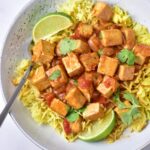 A bowl of yellow rice topped with creamy vegan tofu curry in a tomato-based sauce, garnished with cilantro and lime wedges, with a spoon on the side.
