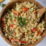 A wooden bowl filled with quinoa zesty salad, featuring chickpeas, diced cucumber, tomato, red onion, and parsley, with a wooden spoon in the bowl.