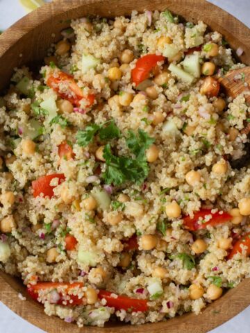 A wooden bowl filled with quinoa zesty salad, featuring chickpeas, diced cucumber, tomato, red onion, and parsley, with a wooden spoon in the bowl.