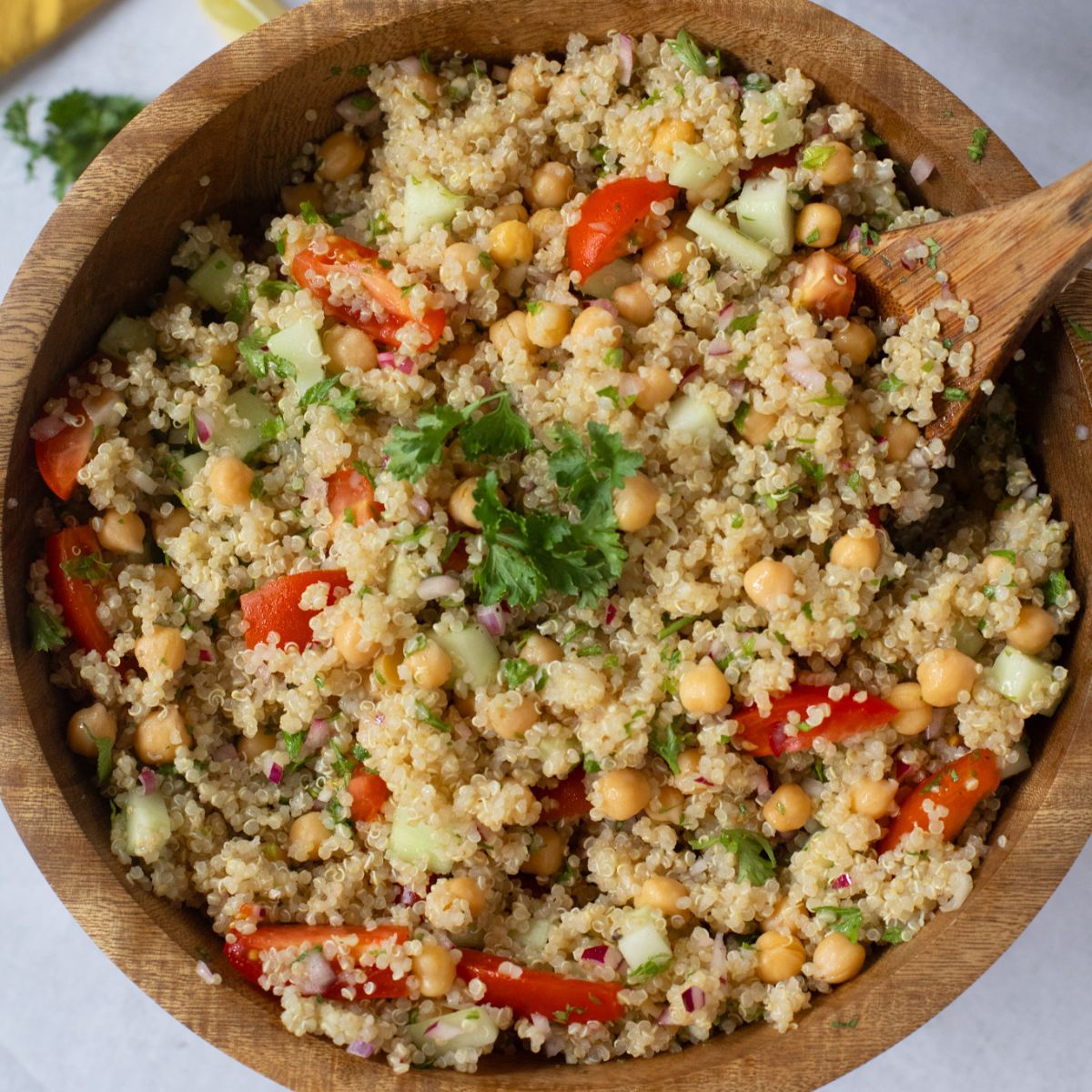 A wooden bowl filled with quinoa zesty salad, featuring chickpeas, diced cucumber, tomato, red onion, and parsley, with a wooden spoon in the bowl.