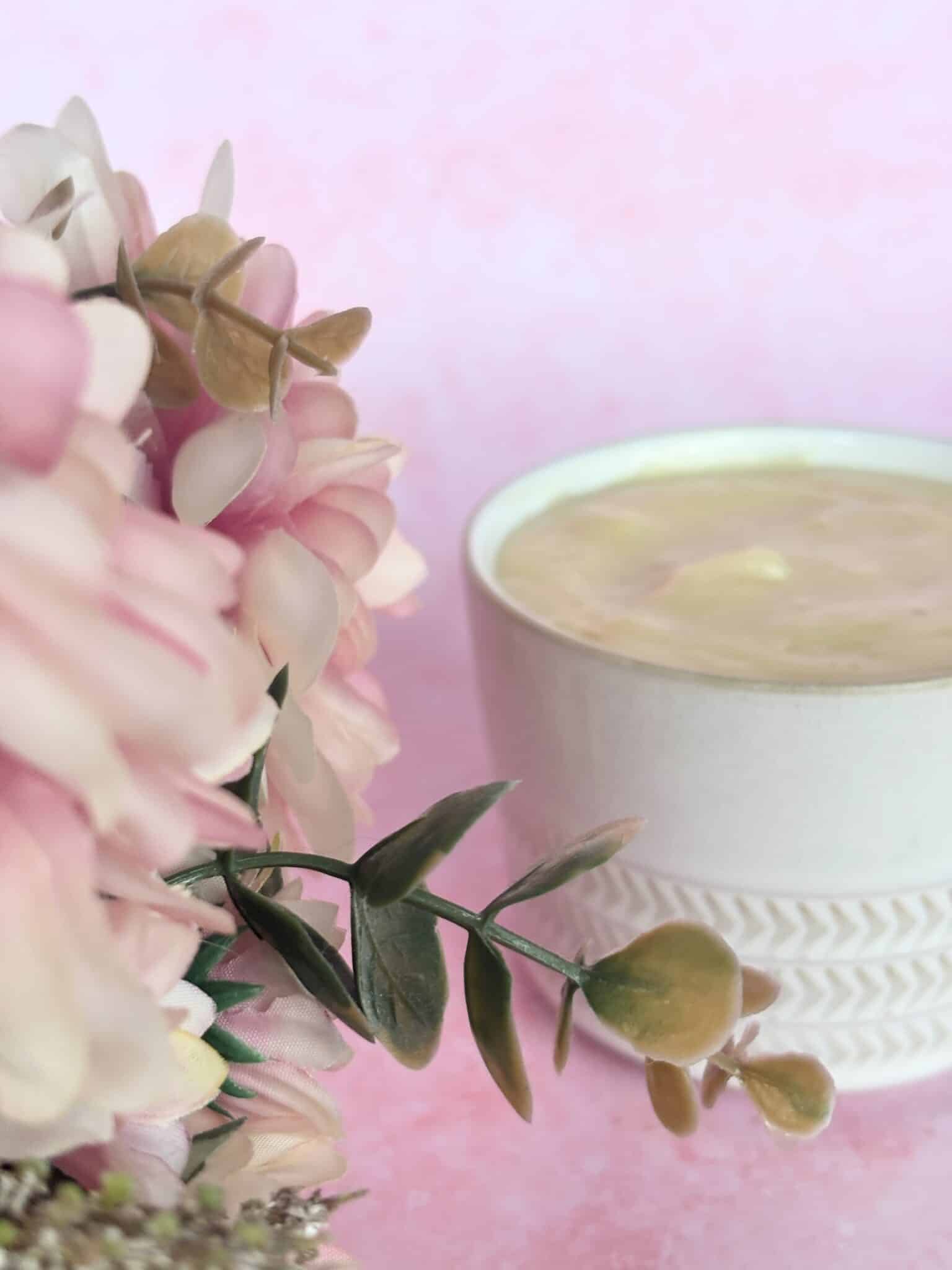 A white ceramic cup filled with a creamy beverage sits next to a bunch of artificial pink flowers against a pink background.