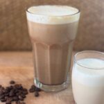 A tall glass of iced coffee with foam next to a small glass of milk and scattered coffee beans on a wooden surface.