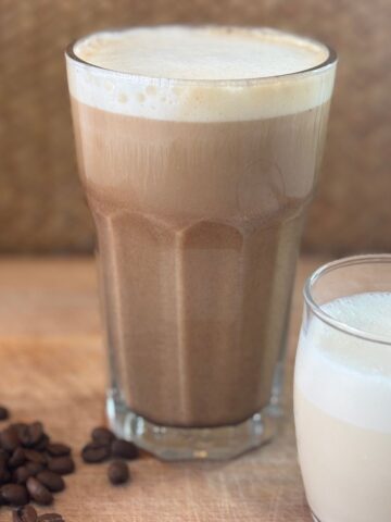 A tall glass of iced coffee with foam next to a small glass of milk and scattered coffee beans on a wooden surface.