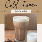 A glass of iced coffee topped with vegan cold foam sits next to a smaller glass of foam, with coffee beans scattered on the table.