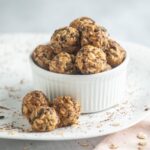 A white ramekin filled with vegan peanut butter balls, oat, and chocolate chip energy bites, with a few more bites and oat flakes scattered on a white plate.