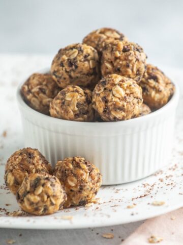 A white ramekin filled with vegan peanut butter balls, oat, and chocolate chip energy bites, with a few more bites and oat flakes scattered on a white plate.