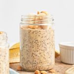 A glass jar filled with Vegan Overnight Oats chia seed pudding, topped with banana slices and peanuts, sits on a wooden surface next to a cut banana and a small white dish.