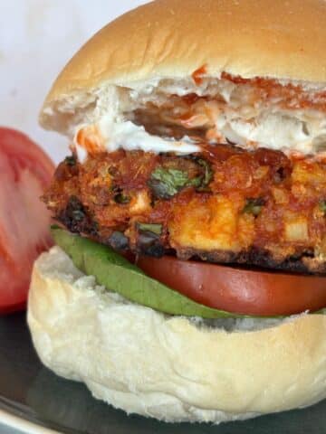 A close-up of a veggie burger with lettuce, tomato, and sauce in a bun, served on a plate. A sliced tomato is placed next to the burger.