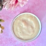 A bowl filled with a creamy, light beige substance is placed on a pink surface, with pink flowers partially visible at the top left.