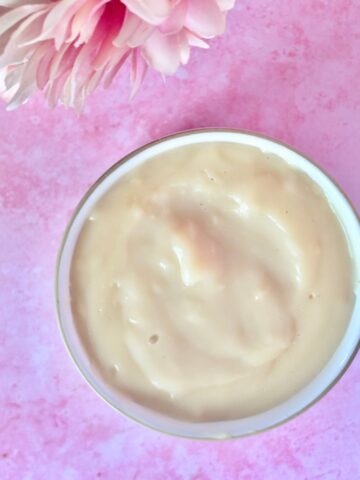 A bowl filled with a creamy, light beige substance is placed on a pink surface, with pink flowers partially visible at the top left.