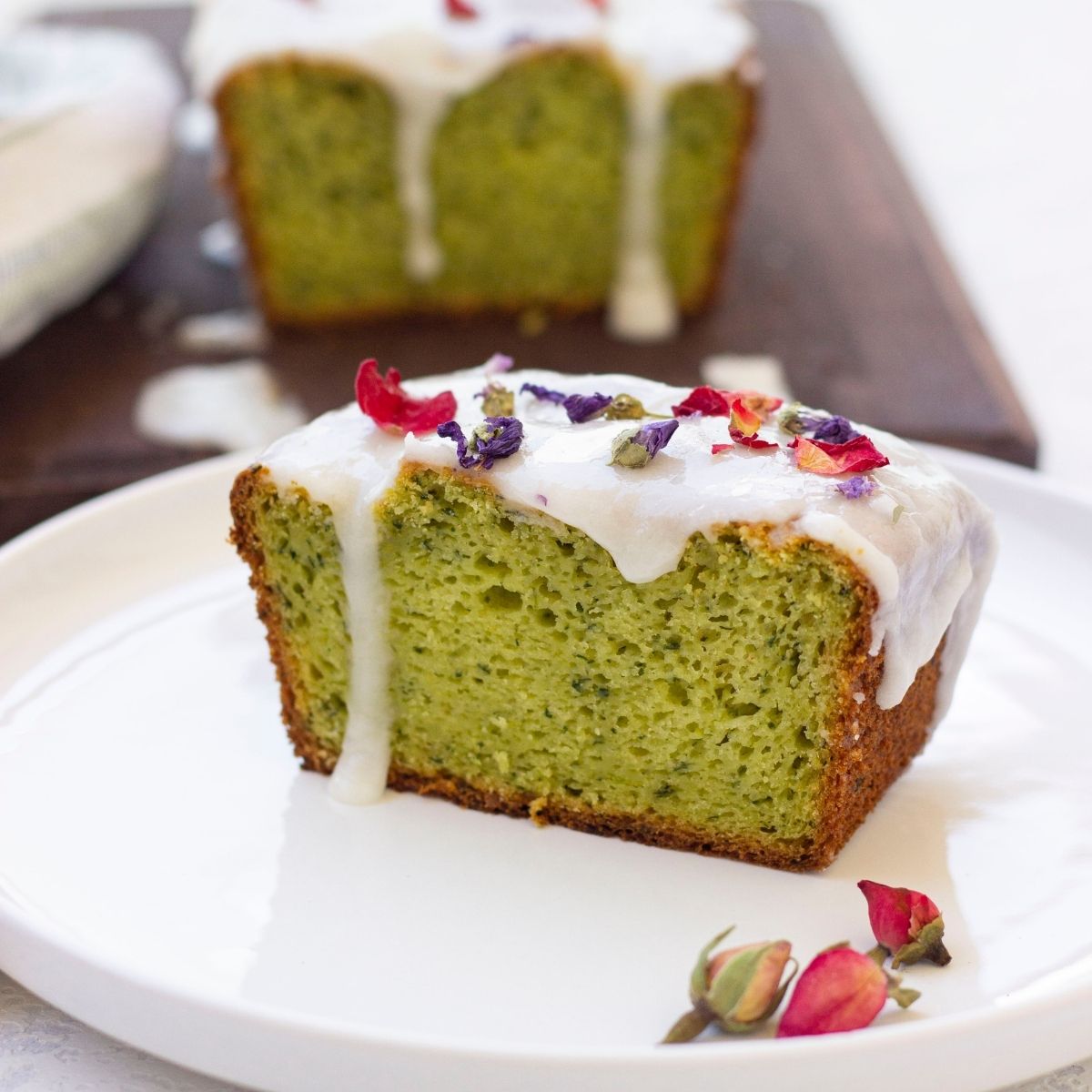 A slice of Vegan Avocado Cake with lemon icing and dried flower petals rests on a white plate, while the rest of this delightful vegan cake recipe sits in the background.
