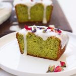 A slice of vegan avocado cake with lemon icing and dried flower petals on top, served on a white plate. The rest of the loaf is in the background.