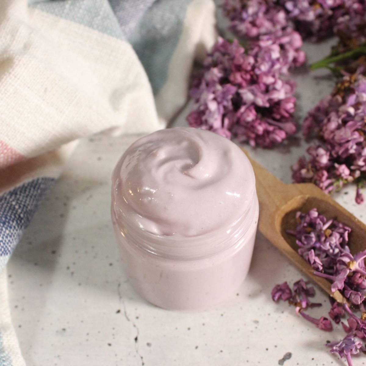 A small glass jar filled with light purple cream sits on a white surface, surrounded by lilac flowers and a wooden scoop.