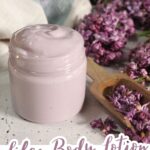 A small glass jar filled with light purple lotion sits on a white surface next to dried lilac flowers and a wooden scoop. The label reads "Lilac Body Lotion: Natural and homemade.
