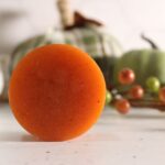 An orange pumpkin spice soap bar stands upright on a white surface, with blurred pumpkins and berry decorations in the background.