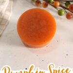 Round orange bar of homemade soap sits on a white surface, surrounded by a fall-themed arrangement and a plaid cloth. Text reads “Pumpkin Spice Scented Homemade Soap,” featuring an easy soap recipe perfect for DIY Pumpkin Spice Soap lovers.
