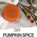 Round orange pumpkin spice soap on a checkered cloth, next to decorative artificial berries and textiles. Text overlay reads "DIY Pumpkin Spice Soap." Try this easy soap recipe for a cozy touch—perfect for anyone seeking a natural soap recipe.