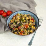A bowl of high protein quinoa salad with black beans, corn, cherry tomatoes, and herbs sits next to a fork and a few loose tomatoes on a light surface.