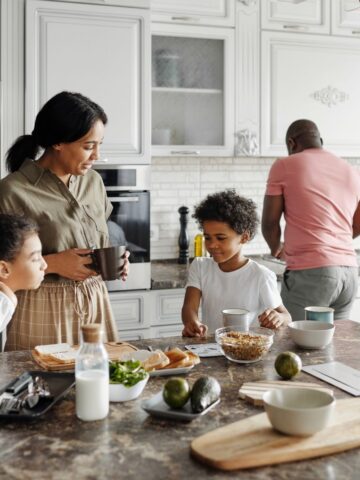 Lazy Plant Based Dinner Ideas For Family Mealtimes 15 A woman and two children stand at a kitchen island with breakfast items, while a man prepares food at the stove in the background.