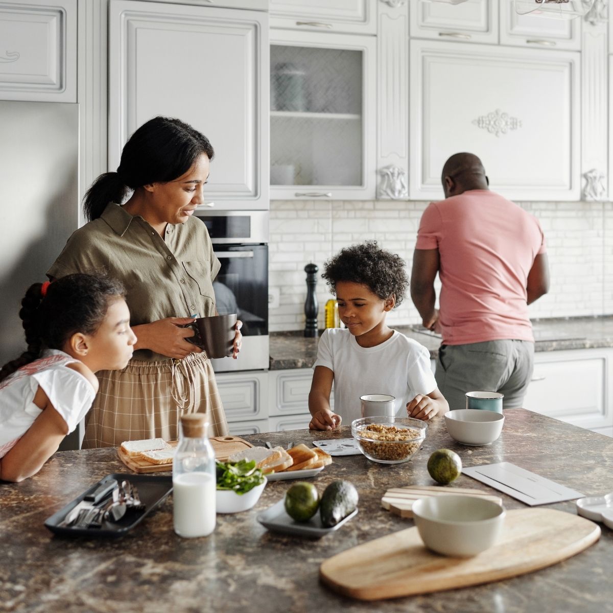 A woman and two children stand at a kitchen island with breakfast items, while a man prepares food at the stove in the background.