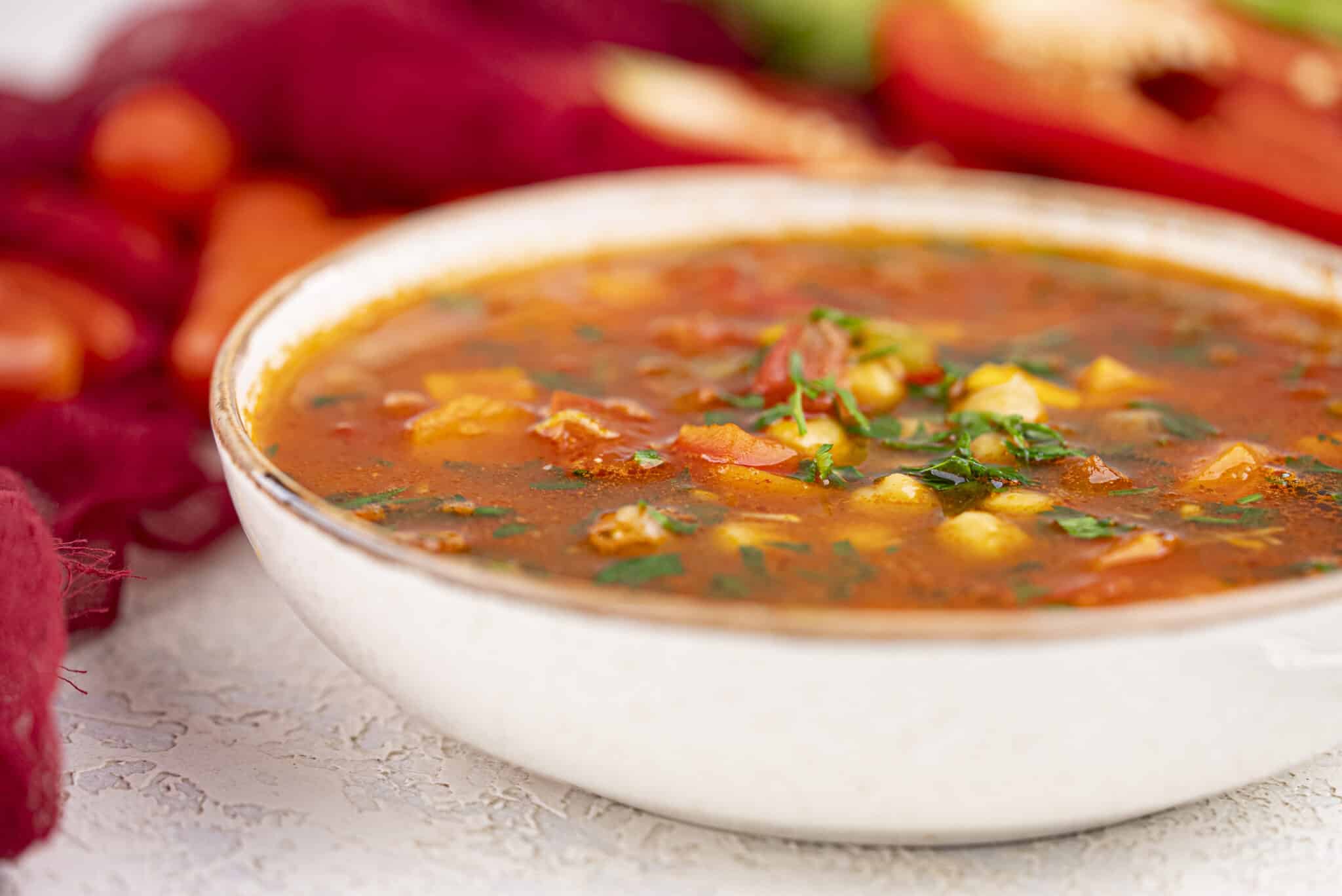 A white bowl filled with vegetable soup, featuring chunks of vegetables and fresh herbs, sits on a light-colored surface with blurred produce in the background.