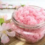 A small glass jar filled with Cherry Scrub for body exfoliation sits on a wooden surface next to light pink artificial flowers.