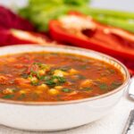 A bowl of vegan vegetable soup with diced vegetables in a red broth, featuring fresh celery and red bell pepper in the background.