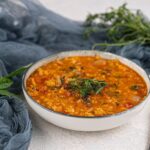 A bowl of thick vegan lentil soup garnished with fresh herbs, placed on a light textured surface with greens and fabric in the background—a comforting slow cooker dish perfect for cozy meals.