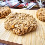 A close-up of a vegan oatmeal cookie on a wooden board, with more oatmeal cookies and a plaid cloth in the background.