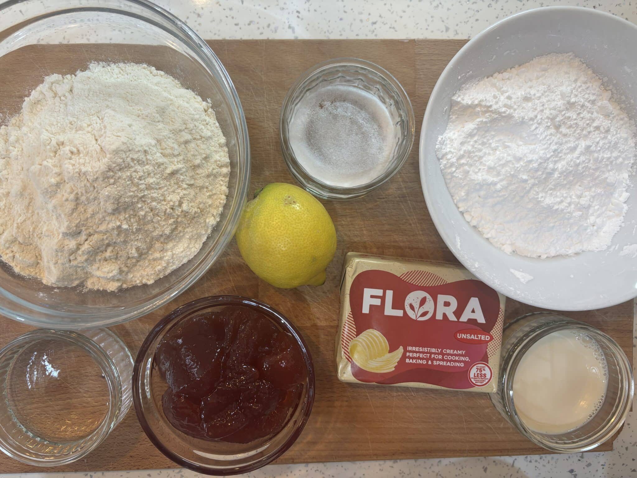 Vegan Pop Tart Recipe (Homemade Strawberry Pop-Tarts) 261 A wooden board displays flour, powdered sugar, jam, a lemon, vegan margarine, sugar, water, and plant milk in bowls and containers, arranged for baking preparation.
