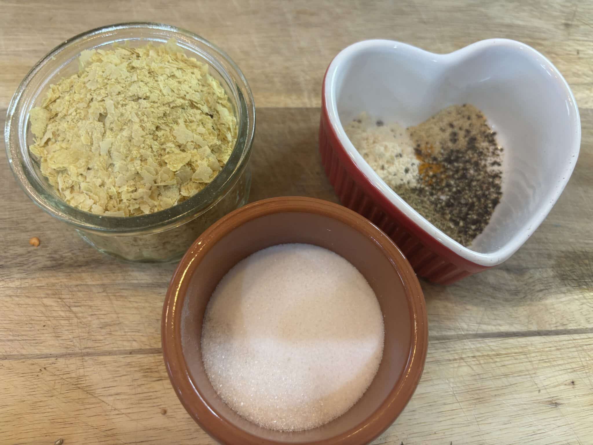 Vegan Chicken Broth Recipe (Homemade Bouillon Powder) 183 Three small containers with ingredients: one with granulated white sugar, one with yellow nutritional yeast flakes, and one with mixed spices, on a wooden surface.