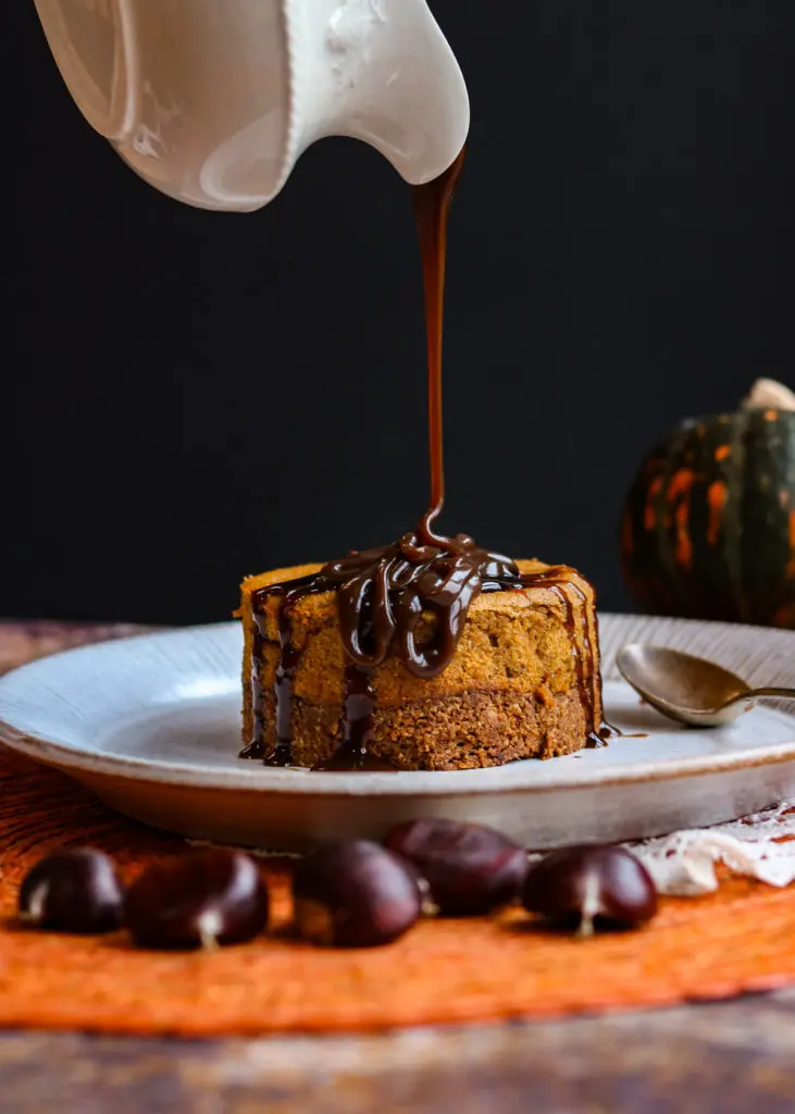 Vegan Thanksgiving Desserts - Go-To Best Dessert Recipes 16 Chocolate sauce is being poured from a white pitcher onto a slice of cake on a plate, with chestnuts and a pumpkin in the background-perfect inspiration for Vegan Thanksgiving Desserts or anyone seeking the Best Dessert Recipes.