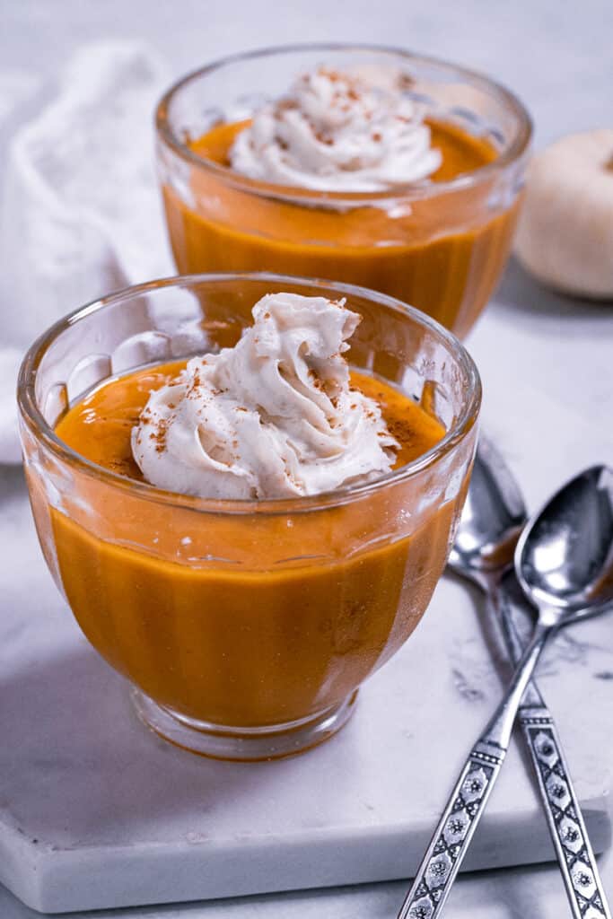 Vegan Thanksgiving Desserts - Go-To Best Dessert Recipes 23 Two glass bowls filled with orange pumpkin pudding, each topped with whipped cream and a sprinkle of spice, sit on a marble surface with two spoons-perfect for trying the best dessert recipes or adding to your vegan Thanksgiving desserts spread.