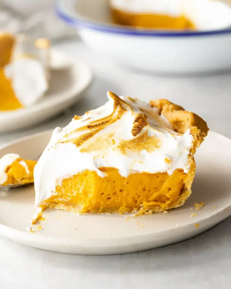 Vegan Thanksgiving Desserts - Go-To Best Dessert Recipes 21 A slice of pumpkin pie topped with toasted meringue sits on a white plate-an inviting addition to any Vegan Thanksgiving Desserts spread, with a serving dish and fork in the background.