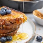 Three slices of cinnamon sugar toast stacked on a plate, topped with blueberries and drizzled with honey. A bowl of cinnamon and a skillet are in the background.