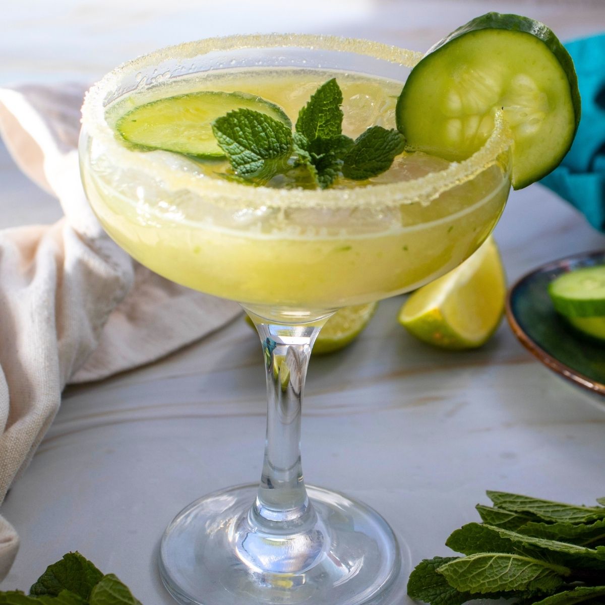 A margarita in a salt-rimmed glass, garnished with cucumber slices and fresh mint, sits on a light surface with lime wedges and mint nearby—perfect inspiration for a refreshing cucumber mocktail.