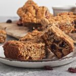 Close-up of two oat and nut bars with visible dried fruit on a ceramic plate, with more bars and a small bowl in the background.