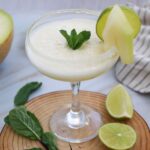 A cocktail glass with a light yellow frozen Margarita Mocktail, garnished with fresh mint, a lime wheel, and melon slices, sits on a wooden coaster beside lime halves and mint leaves—an easy recipe perfect for summer melon lovers.