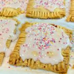 Rectangular pastries with light pink icing and colorful sprinkles on top, arranged on parchment paper—these treats are inspired by a delicious vegan pop tart recipe.