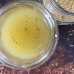 A glass jar filled with vibrant yellow Vegan Chicken Broth, seasoned with Homemade Bouillon Powder, sits on a dark brown surface near a partially visible jar of dried flakes.