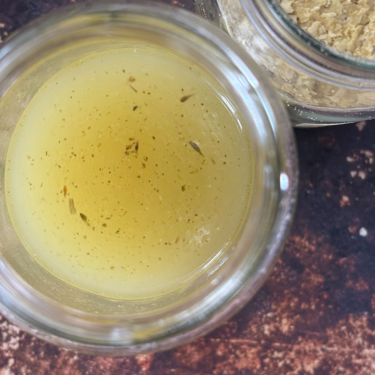 A glass jar filled with vibrant yellow Vegan Chicken Broth, seasoned with Homemade Bouillon Powder, sits on a dark brown surface near a partially visible jar of dried flakes.