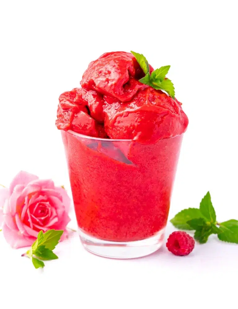 50 Delicious Vegan Desserts With Fruit That You Will Love 15 A glass filled with bright red raspberry sorbet, one of the finest vegan desserts with fruit, is garnished with mint leaves. A pink rose, mint sprigs, and a raspberry sit beside the glass on a white background.