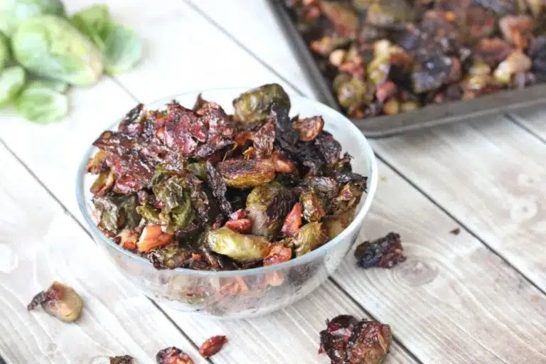 Family-Pleasing Easy Vegan Thanksgiving Recipes 13 A glass bowl filled with roasted Brussels sprouts and pecans sits on a wooden surface, with a baking tray of more roasted vegetables in the background.