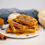 A stack of fluffy Vegan Pumpkin Waffles drizzled with syrup on a white plate, with a cinnamon stick, pumpkin seeds, and white pumpkins in the background.