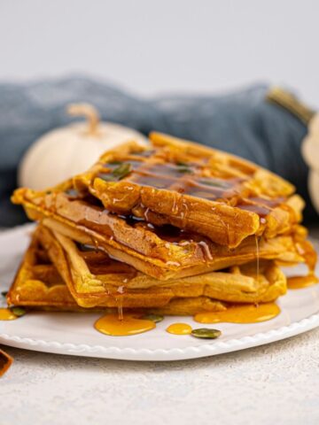 A stack of fluffy Vegan Pumpkin Waffles drizzled with syrup on a white plate, with a cinnamon stick, pumpkin seeds, and white pumpkins in the background.